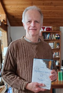 Michael Dolzani standing in a living room holding a copy of his book, The Productions of Time.